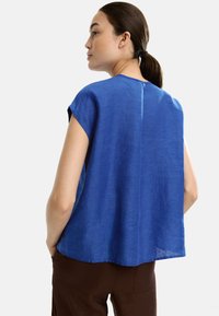Niebieska lniana bluzka z krótkimi rękawami, z tylnym szwem i luźnym krojem; noszona z brązowymi spodniami.
