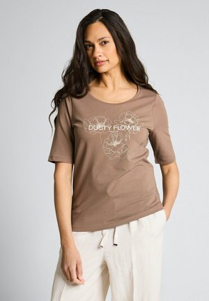 Kobieta o długich ciemnych włosach, ubrana w brązowy T-shirt "Dusty Flower" oraz białe spodnie ze ściągaczem, stojąca na jednolitym jasnoszarym tle.