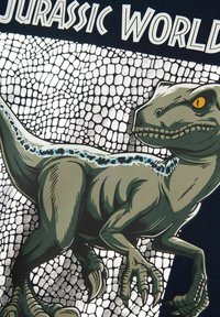 T-Shirt mit einem Grafik eines grünen Dinosauriers mit einem orangenen Auge, vor einem netzartigen Hintergrund und dem Text "Jurassic World."