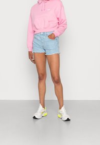 Růžová krátká mikina s přední kapsou, v kombinaci se světle modrými džínovými šortkami s třepícím se lemem a jemnými vertikálními pruhy, a bílými teniskami.