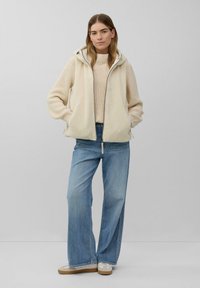 Frau steht mit Händen in den Taschen, trägt einen cremefarbenen Strickpullover, eine cremefarbene Fleeceweste mit Kapuze, weit geschnittene blaue Jeans und weiße Sneakers vor einem grauen Hintergrund.