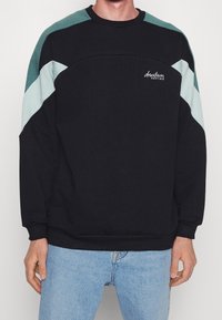 Sudadera negra con diseño geométrico en bloque de color verde azulado y azul claro. Presenta cuello redondo y puños acanalados. Logo en el pecho.