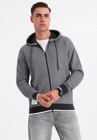 Sudadera gris con cremallera y detalles en negro, con cordones y puños acanalados. Tejido suave con un dobladillo recto, llevada sobre una camiseta blanca y jeans claros.