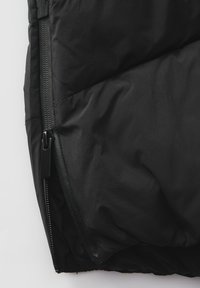 Schwarzer gepolsterter Jackenteil mit sichtbarem Reißverschluss, der eine glatte Textur und gestickte Paneele aufweist. Der Reißverschlusszug ist matt schwarz.