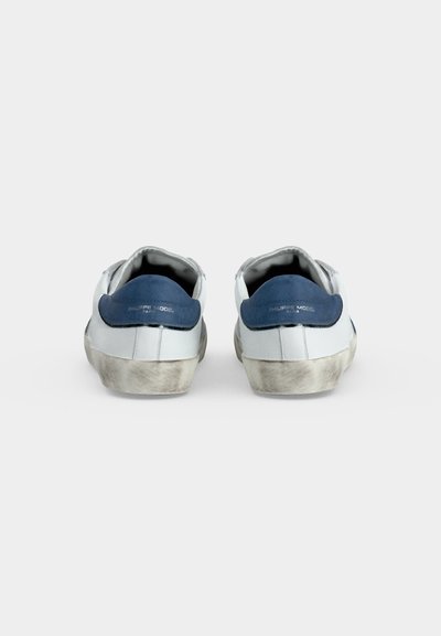 Paire de baskets blanches basses avec languettes de talon bleu marine et semelles usées et sales, vue de dos sur fond uni.