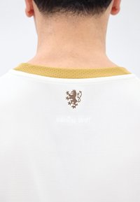 Οπίσθια όψη ενός ατόμου που φοράει λευκό πουκάμισο με χρυσό ριγωτό γιακά και μικρό καφέ έμβλημα λιονταριού κάτω από το λαιμό.