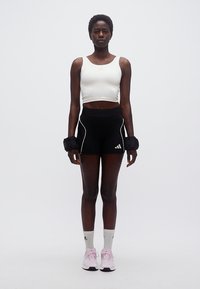 Γυναίκα όρθια, φορώντας λευκό crop top, μαύρο σορτς, βαράκια καρπού, λευκές κάλτσες και ροζ αθλητικά παπούτσια, μπροστά σε απλό φόντο.