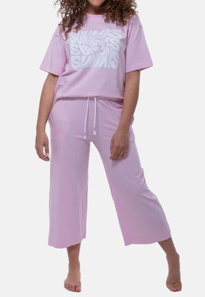 Vrouw in roze losse broek en bijpassend kort mouwshirt met witte draaiende "SUNDAYS" tekst, staat op blote voeten tegen een witte achtergrond.