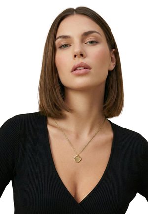 Donna con capelli castani lisci che indossa una maglia nera a scollo a V e una collana con ciondolo circolare dorato su una fine catena.
