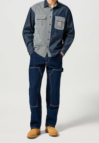 Džínová košile se pruhovanými a jednobarevnými tmavě modrými panely, dvěma předními kapsami; spárovaná s modrými džínami a hnědými pracovními botami.