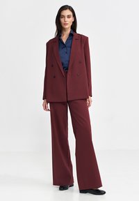 Burgundowa dwurzędowa marynarka i pasujące szerokie spodnie, w połączeniu z ciemnoniebieską koszulą. Gładki materiał z minimalnymi detalami, dopasowany krój.