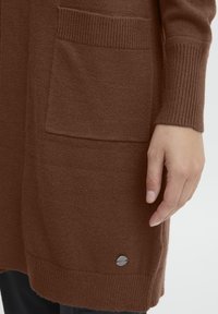 Bruine gebreide cardigan met geribde mouwen en twee voorzakken. Een klein metalen logo-detail is zichtbaar nabij de ondernaad.