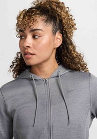 Pilkas zip-up gobtuvas, pagamintas iš tekstūruoto audinio, su pritaikytu dizainu. Yra raišteliai, Nike logotipas ir gobtuvas. Matomi garbanoti plaukai.