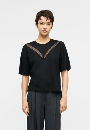 Jong persoon met kort haar draagt een zwart topje met V-vormig kantdetail en losse donkergrijze broek, staand tegen een effen achtergrond.