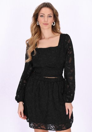 Mladá žena s dlouhými vlnitými vlasy, která nosí černé průsvitné texturované šaty s dlouhými rukávy a čtvercovým výstřihem, doplněné kruhovými náušnicemi.