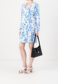 Gaun floral berwarna biru muda dan putih dengan lengan panjang, leher V yang dalam, dan desain bertingkat; dipadukan dengan tas tangan hitam dan sandal bertumit blok putih.