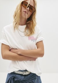 En blond person iført oversized amber solbriller, en hvid t-shirt med pink tekst, lagdelte sølvkæder og denim bukser bundet med et nittebesat hvidt bælte.