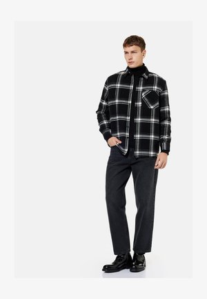 Joven de pie con camisa de cuadros en blanco y negro sobre un jersey de cuello alto negro, vaqueros negros y botas negras pulidas sobre fondo blanco.