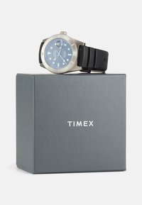 Montre Timex avec un cadran bleu, un boîtier en acier inoxydable et un bracelet en silicone noir sur une boîte grise. Les caractéristiques notables incluent des aiguilles et des marqueurs lumineux.