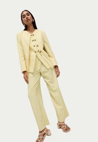 Yellow tailored suit featuring a structured jacket with five circular buttons and straight-leg pants. Paired with beige strappy heeled sandals.