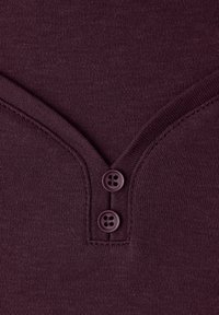 Top in maglia color bordeaux con scollo a V e due bottoni sul collo. Il tessuto ha una texture liscia con dettagli a coste lungo i bordi.