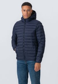 Marineblå pufferjakke med vandret quiltedesign, lynlås foran og hætte. Båret over denimjeans, der fremhæver en glat, let tekstur.