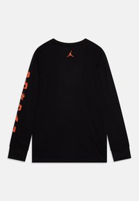 Camiseta negra de manga larga con un logo Jumpman naranja en la parte posterior y un gráfico de texto naranja a lo largo de la manga izquierda. Fabricada en suave algodón.