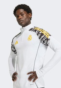 White long-sleeve athletic top with half-zip, black and gray patterned shoulders, yellow logo, and side panel accents. Made from moisture-wicking material.