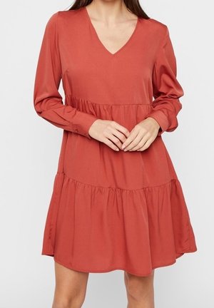 Vrouw draagt een roestkleurige mini-jurk met lange mouwen, V-hals, met lagen en plooien die voor een losse pasvorm zorgen.