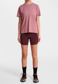 Rosa Baumwoll-T-Shirt mit einem einfachen Logo, kombiniert mit kastanienbraunen Radshorts. Schwarze Kniestrümpfe und mehrfarbige Sportschuhe runden das Outfit ab.