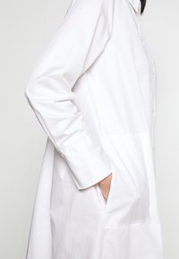 Persona che indossa una lunga camicia bianca con bottoni, con le maniche abbassate e la mano inserita nella tasca laterale, su uno sfondo semplice.