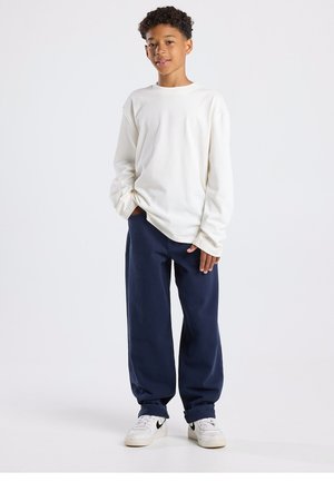 Camisa blanca de algodón de manga larga combinada con pantalones anchos azul marino oscuro y zapatillas blancas con detalles en negro. Corte casual con silueta relajada.