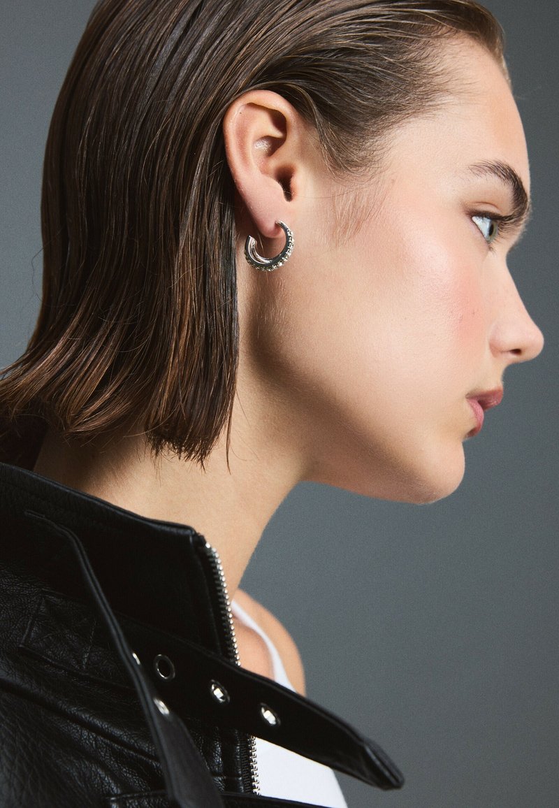 Jeune femme aux cheveux courts et bruns portant une boucle d’oreille ronde en argent et une veste en cuir noire, vue de profil sur un fond gris.