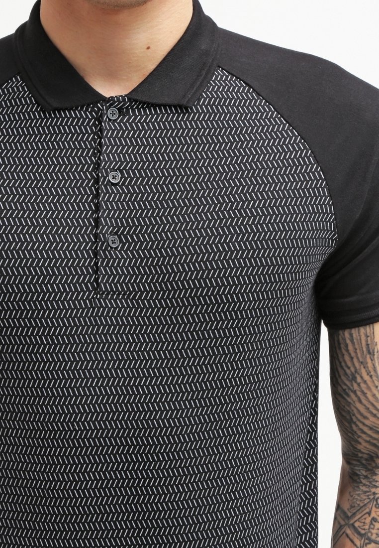 Homme portant un polo noir avec un motif de tirets diagonaux blancs et des manches raglan noires unies, montrant son avant-bras tatoué.