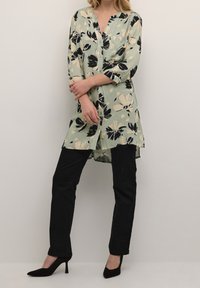 Tunique à boutons florale en vert menthe avec des fleurs noires et crème, associée à un pantalon noir et des chaussures à talons hauts. Encolure simple, manches trois-quarts.