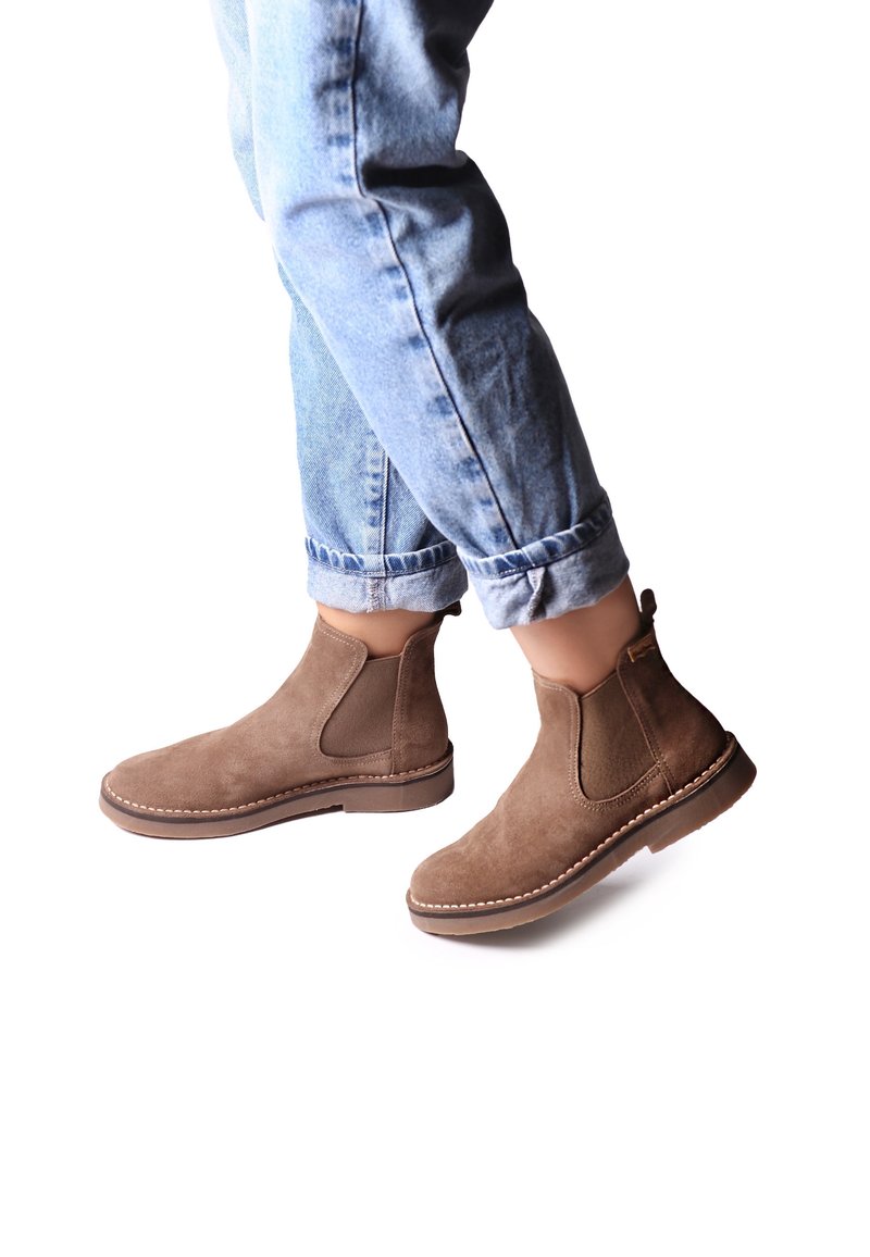 Jambes portant un jean bleu clair retroussé et des bottines marron en daim avec des panneaux latéraux élastiques sur fond blanc.