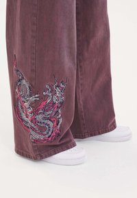 Breda denimbyxor i mörk mauve med färgglad drakbroderi på vänster ben, i kombination med vita sneakers.