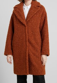 Brun texturerad kappa med en fluffig utseende, med en spetsig krage och öppen framsidan, visad på en person som bär en vit skjorta.