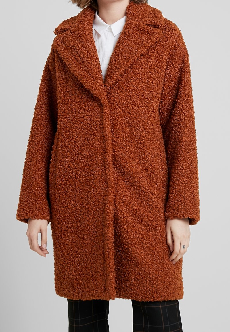 Brun texturerad kappa med en fluffig utseende, med en spetsig krage och öppen framsidan, visad på en person som bär en vit skjorta.