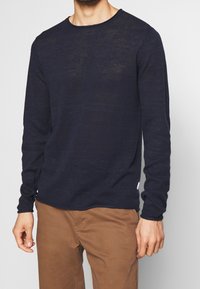 Muž v černém dlouhém svetr s dlouhým rukávem z lehkého pleteného materiálu a hnědých běžných kalhotách, stojící na jednobarevném pozadí.