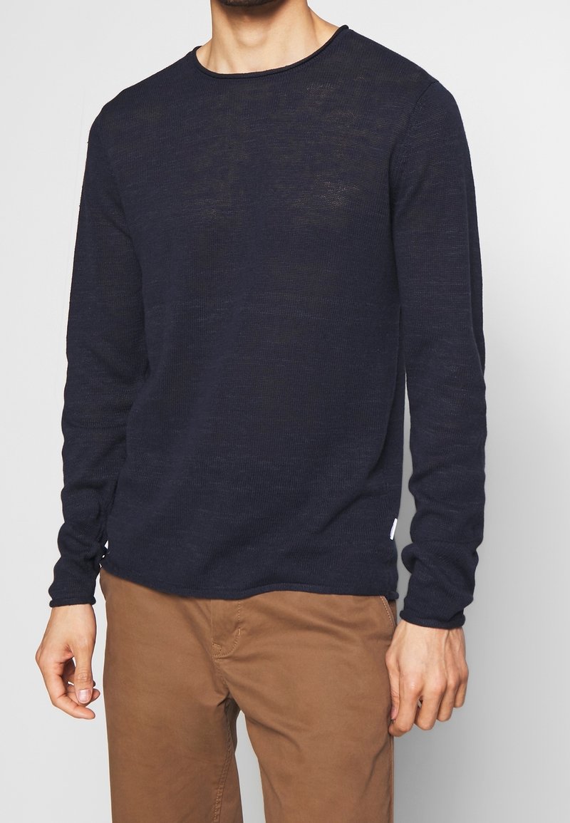 Muž v černém dlouhém svetr s dlouhým rukávem z lehkého pleteného materiálu a hnědých běžných kalhotách, stojící na jednobarevném pozadí.