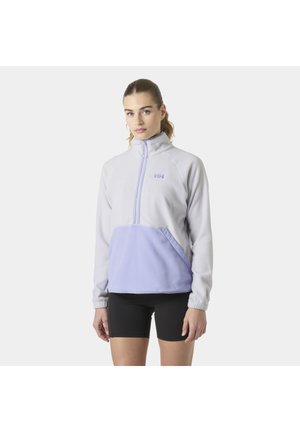 Mujer vistiendo un forro polar gris claro y lavanda con cierre de cuarto y logo, combinado con pantalones cortos de ciclismo negros, de pie frente a un fondo liso.