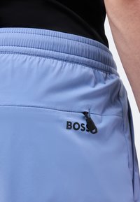 Ανοιχτό μπλε ελαστικό σορτς με μαύρη τσέπη με φερμουάρ και λογότυπο "BOSS" στο πλάι, φορεμένα με μαύρο τοπ.