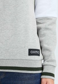Grijze sweatshirt met een witte streep op de mouw, voorzien van een geribbelde groene zoom en boorden, en een zwart "oxmo" logo patch op de taille.