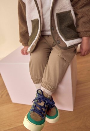 Pantalones de puño elástico color beige, calcetines blancos y zapatillas multicolores con acentos verdes y azules. Chaqueta fuzzy beige y marrón con bolsillos.