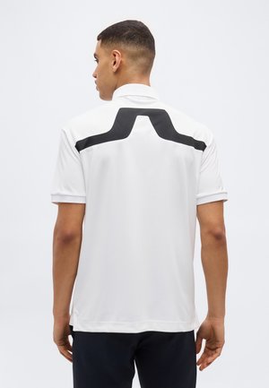 Mand set bagfra iført hvid kortærmet polo med sort geometrisk mønster over øvre ryg mod ensfarvet baggrund.