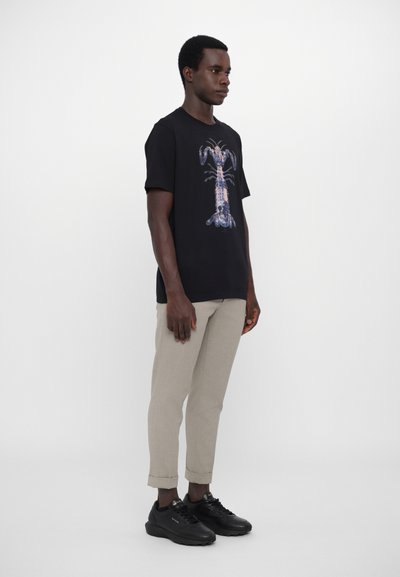 T-shirt noir avec un graphique de homard coloré, associé à un pantalon beige clair et des baskets noires. Le tissu et la coupe sont décontractés.
