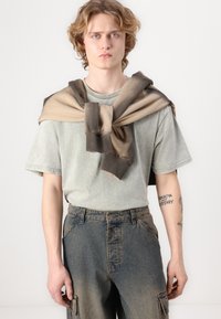 Grau-grünes T-Shirt, über den Schultern getragen mit einem braunen Farbverlauf-Pullover, kombiniert mit blauen schmal geschnittenen Jeans mit Cargo-Taschen.