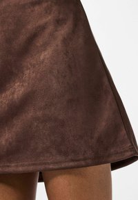 Brun ruskindsundede nederdel med en glat tekstur, udvidet design og synlig syning ved kanten. Stoffet ser blødt og let reflekterende ud.