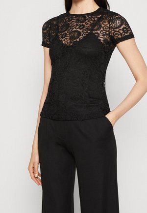 Vrouw draagt een zwart kanten top met korte mouwen met bloemenpatronen en zwarte wijde broek, hand in zak, tegen een effen achtergrond.